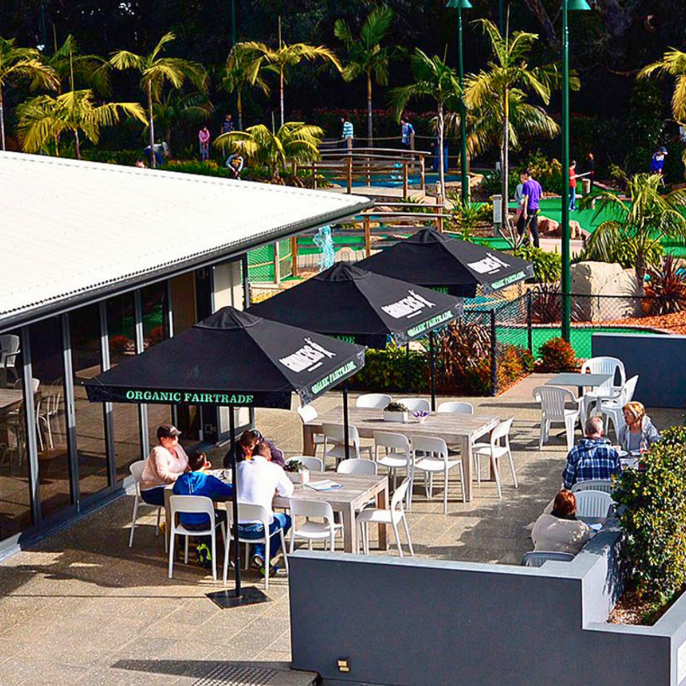 Outdoor patio seating area with multiple black umbrellas over white tables and chairs, with people dining in a sunny cafe setting surrounded by mini golf course